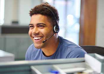 Cheerful customer service representative wearing a headset, smiling as they assist a customer. They are seated in a modern office environmen